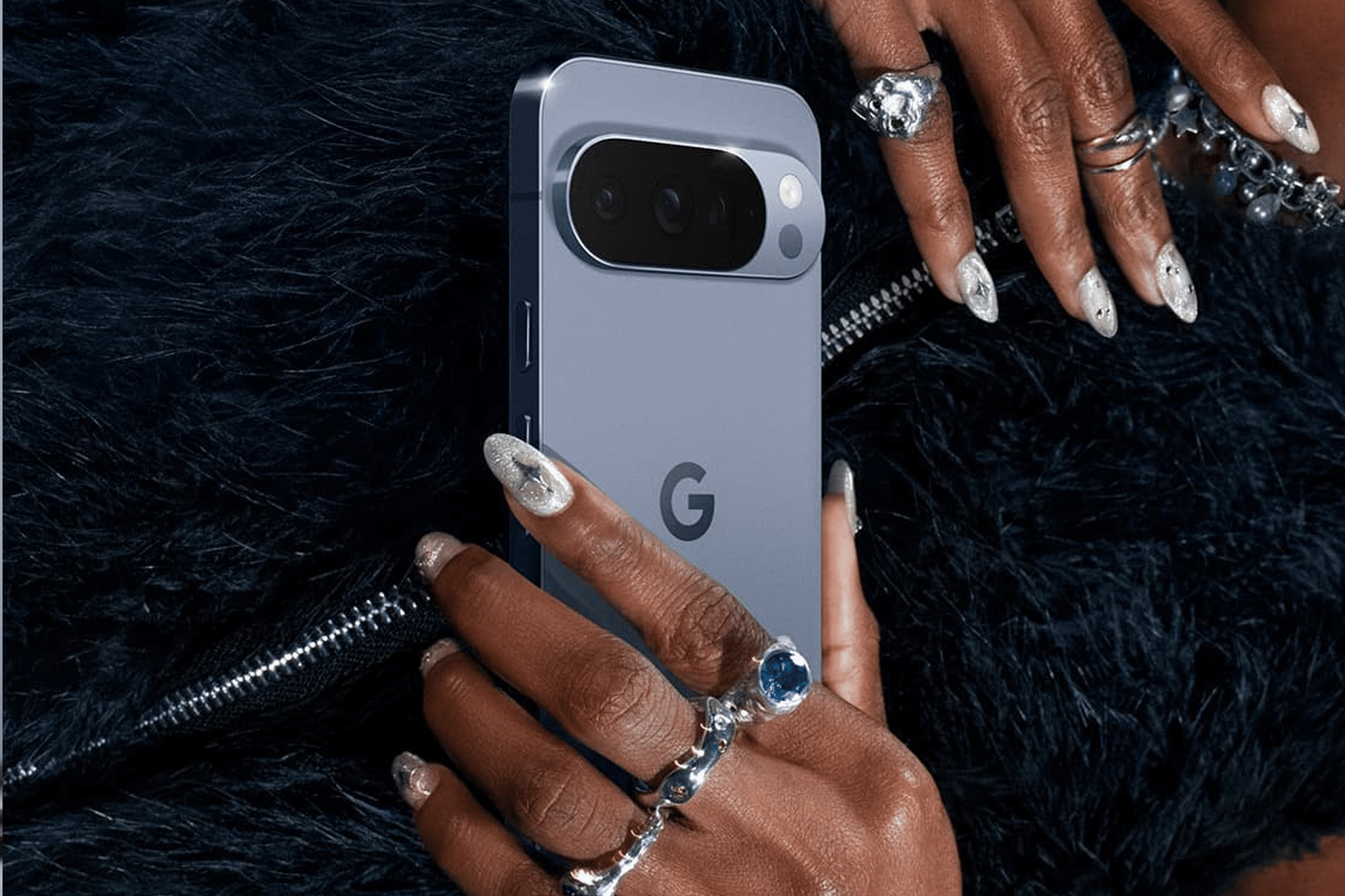 A person with manicured nails and multiple rings holds a blue Google Pixel smartphone against a black, furry textured background—perfect for Pixel users eager to explore the latest Android features.