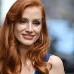 A woman with long wavy red hair, reminiscent of Jessica Chastain, wearing hoop earrings and a blue top, smiles while standing outdoors with a blurred city background.