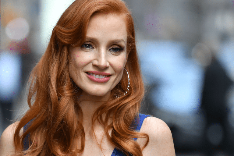 A woman with long wavy red hair, reminiscent of Jessica Chastain, wearing hoop earrings and a blue top, smiles while standing outdoors with a blurred city background.