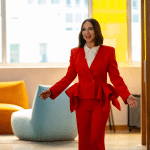 A woman in a bright red suit smiles and walks down a modern office hallway with colorful furniture and large windows—capturing the vibrant energy seen in the Loot Season 3 Trailer on Apple TV Plus.