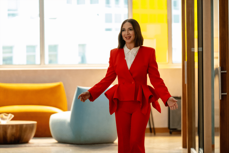 A woman in a bright red suit smiles and walks down a modern office hallway with colorful furniture and large windows—capturing the vibrant energy seen in the Loot Season 3 Trailer on Apple TV Plus.
