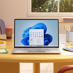 A laptop displaying the Windows 11 start menu sits on a round table surrounded by art supplies, a notebook, and colored pencils in a bright room—perfect for discussing the latest Microsoft Windows features and Windows 12 rumors.