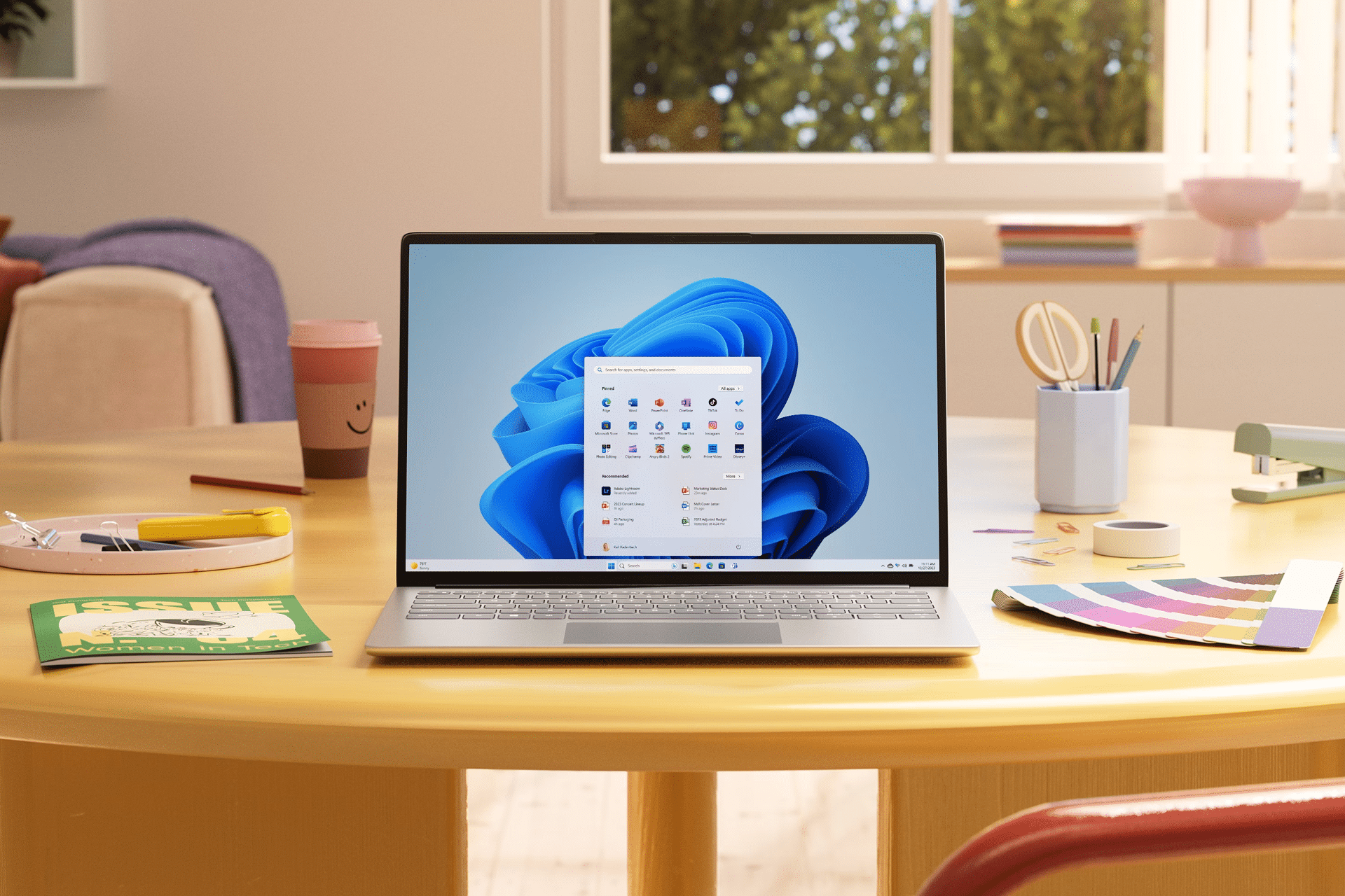 A laptop displaying the Windows 11 start menu sits on a round table surrounded by art supplies, a notebook, and colored pencils in a bright room—perfect for discussing the latest Microsoft Windows features and Windows 12 rumors.