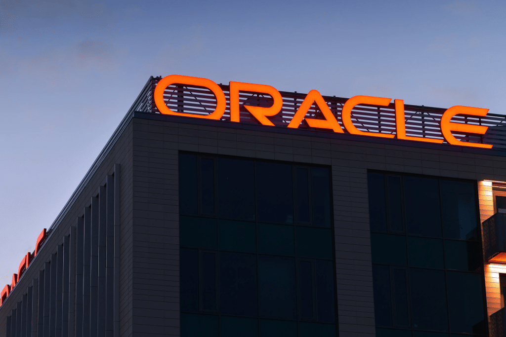 The photo shows the top corner of a modern office building with large, red illuminated letters spelling "Oracle," highlighting its role in AI infrastructure against a dusky sky.
