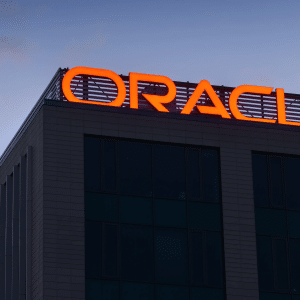 The photo shows the top corner of a modern office building with large, red illuminated letters spelling "Oracle," highlighting its role in AI infrastructure against a dusky sky.
