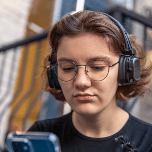 A young person with short brown hair and glasses wears headphones and looks intently at a smartphone, sitting outdoors near a metal railing—benefiting from OpenAI parental safety controls for added peace of mind.