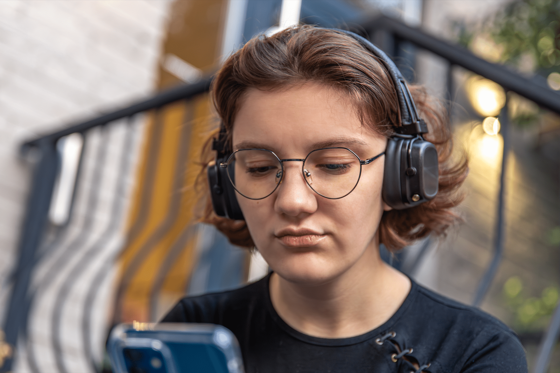 A young person with short brown hair and glasses wears headphones and looks intently at a smartphone, sitting outdoors near a metal railing—benefiting from OpenAI parental safety controls for added peace of mind.
