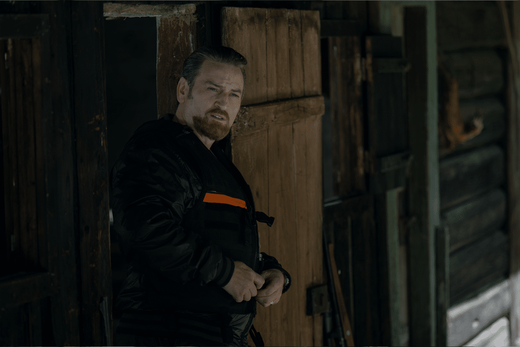 A man with light brown hair and a beard stands in the doorway of a rustic wooden cabin, wearing a black jacket with an orange stripe, echoing the tense mood of Apple TV+ The Hunt Traqués. The cabin is dark with wood-paneled walls and door.