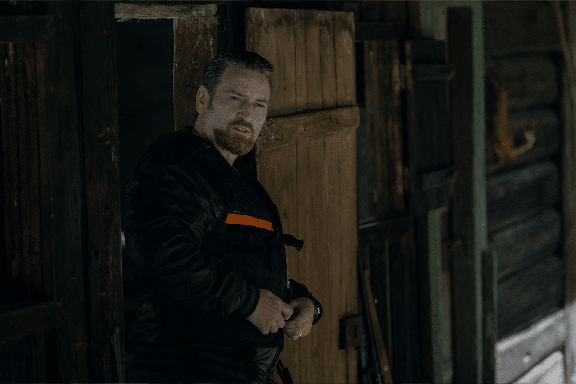A man with light brown hair and a beard stands in the doorway of a rustic wooden cabin, wearing a black jacket with an orange stripe, echoing the tense mood of Apple TV+ The Hunt Traqués. The cabin is dark with wood-paneled walls and door.