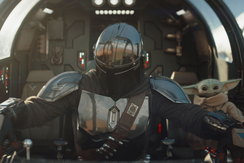 A person in shiny black armor and helmet pilots a spaceship, while The Mandalorian & Grogu sit side by side in the cockpit, surrounded by futuristic controls and lights.