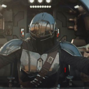 A person in shiny black armor and helmet pilots a spaceship, while The Mandalorian & Grogu sit side by side in the cockpit, surrounded by futuristic controls and lights.