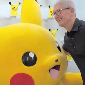 A man in a black shirt smiles while posing next to a large Pikachu mascot, with several smaller Pikachu figures on shelves in the background—capturing a true Tim Cook’s Pikachu Moment. An apple japanese logo is visible in the bottom right corner.