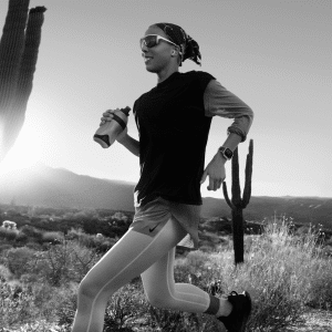 A person jogs through a desert landscape at sunrise, wearing athletic gear, sunglasses, and holding a water bottle. Sporting the Apple Watch 2025, they embrace new features and health innovations as large cacti and mountains rise in the background.