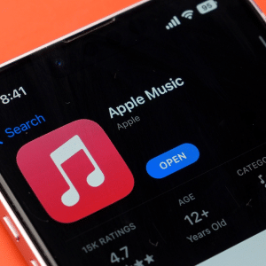 A smartphone screen displays the Apple Music app page in the App Store, featuring the app icon, “Open” button, ratings, age, and category on a bright orange background—now highlighting an iOS 26.1 new gesture for swiping to switch tracks.