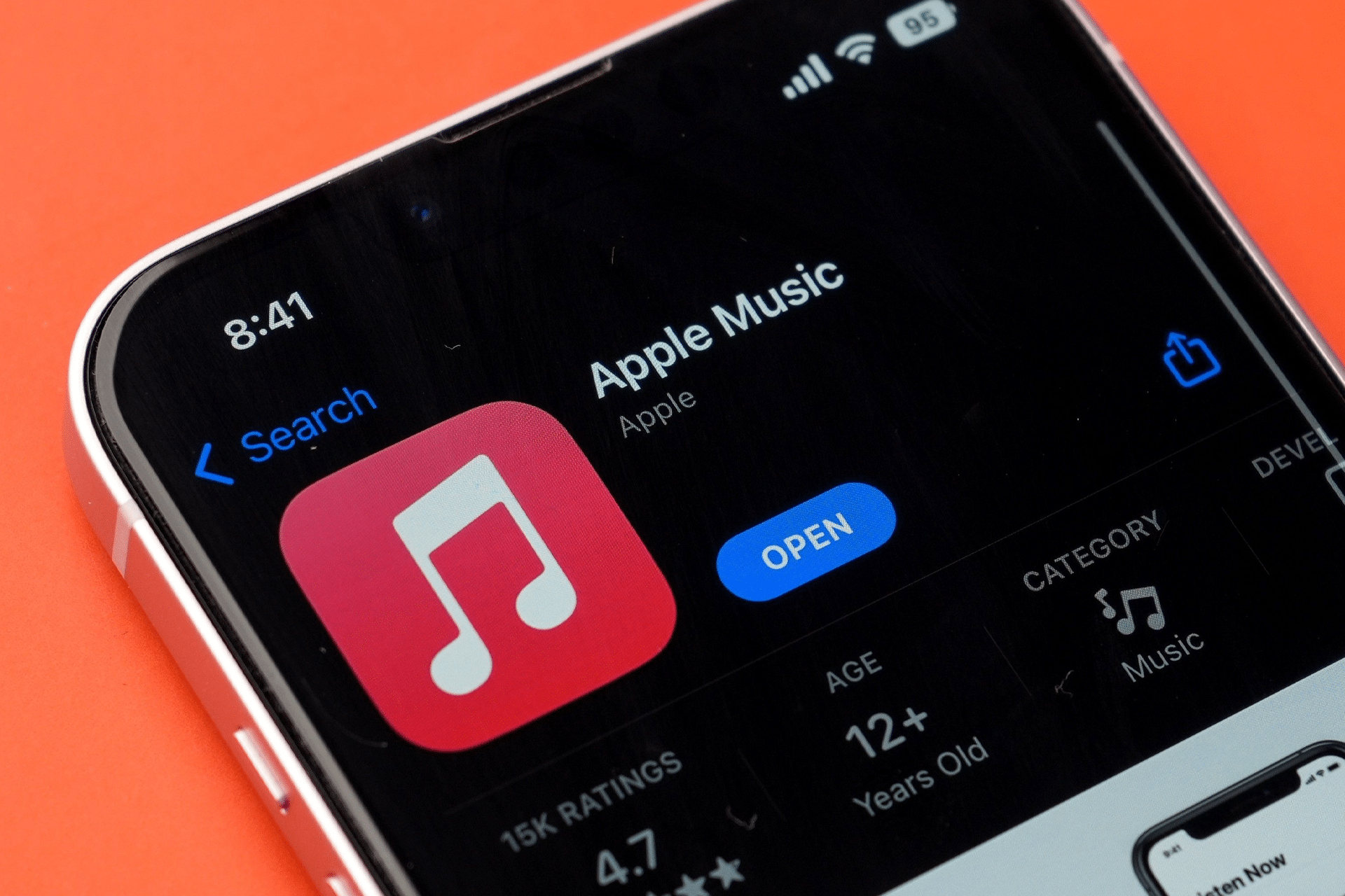 A smartphone screen displays the Apple Music app page in the App Store, featuring the app icon, “Open” button, ratings, age, and category on a bright orange background—now highlighting an iOS 26.1 new gesture for swiping to switch tracks.
