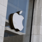 A large, white Apple logo hangs in the window of a modern building with light-colored stone walls and large glass panes, as news emerges of Apple testing chatbot technology inside.