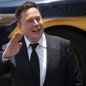 A man in a dark suit and tie smiles and raises his hand in greeting while standing outdoors, with a dark-colored car in the background—like a scene straight out of an Apple TV Plus series.