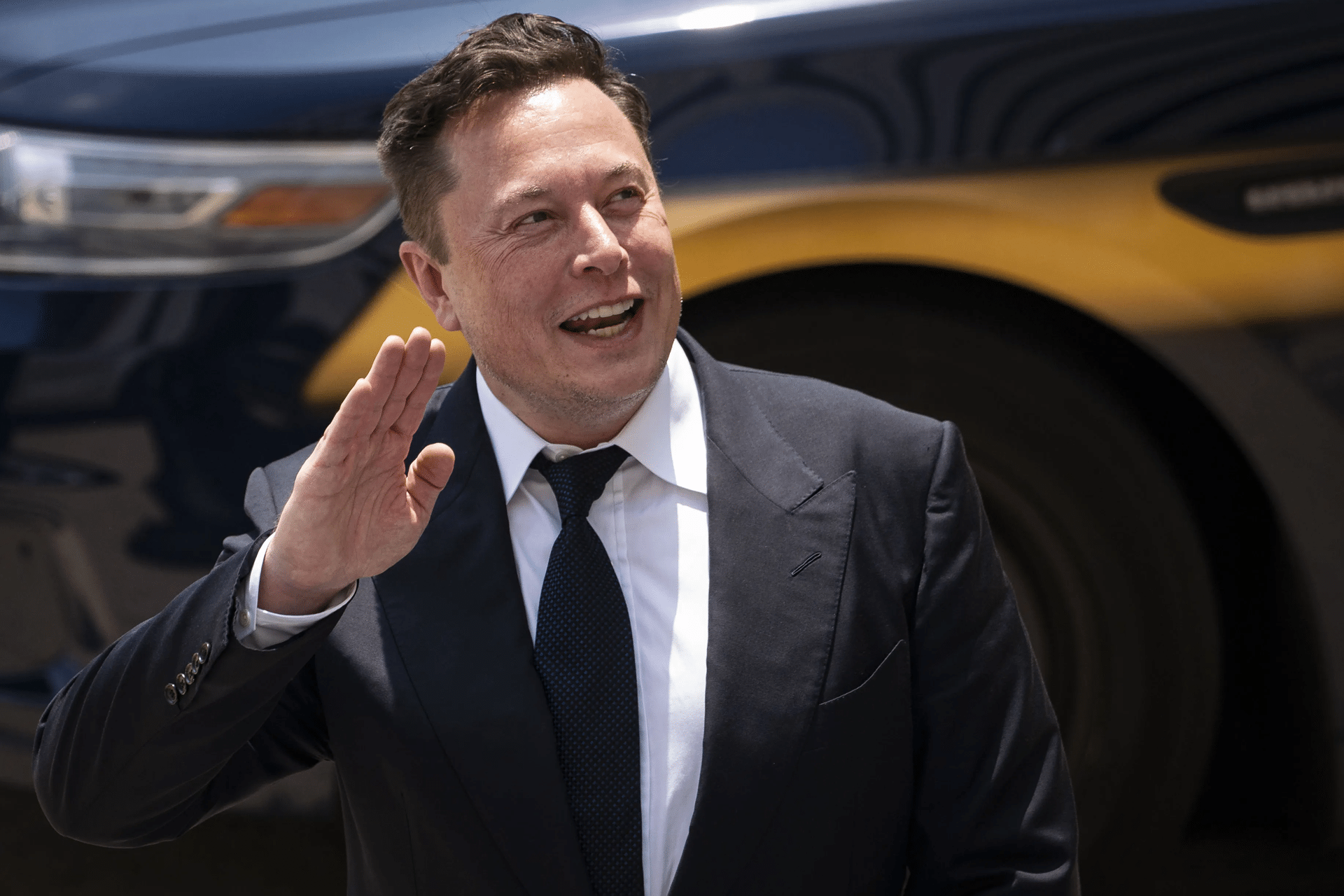 A man in a dark suit and tie smiles and raises his hand in greeting while standing outdoors, with a dark-colored car in the background—like a scene straight out of an Apple TV Plus series.