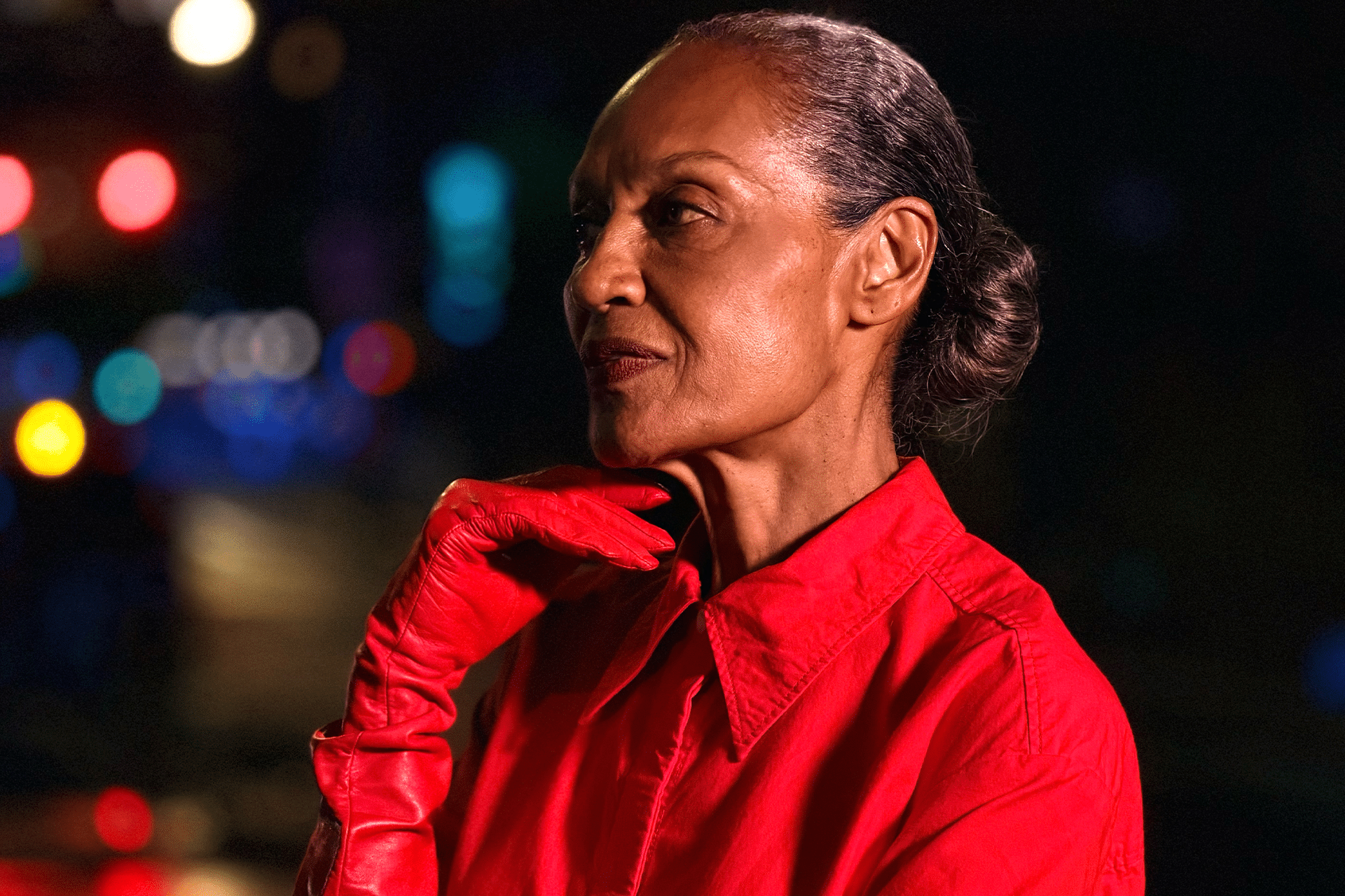 An older woman with dark skin and gray hair pulled back, wearing a bright red shirt and red gloves, poses thoughtfully against a blurred, colorful nighttime background—captured in vivid detail with the iPhone 17 Pro camera system.