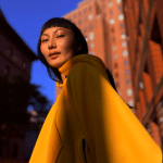 A person wearing a bright yellow coat stands on a city street, partially lit by sunlight, captured using iPhone photography. Red-brick and brown buildings rise in the background beneath a clear blue sky.