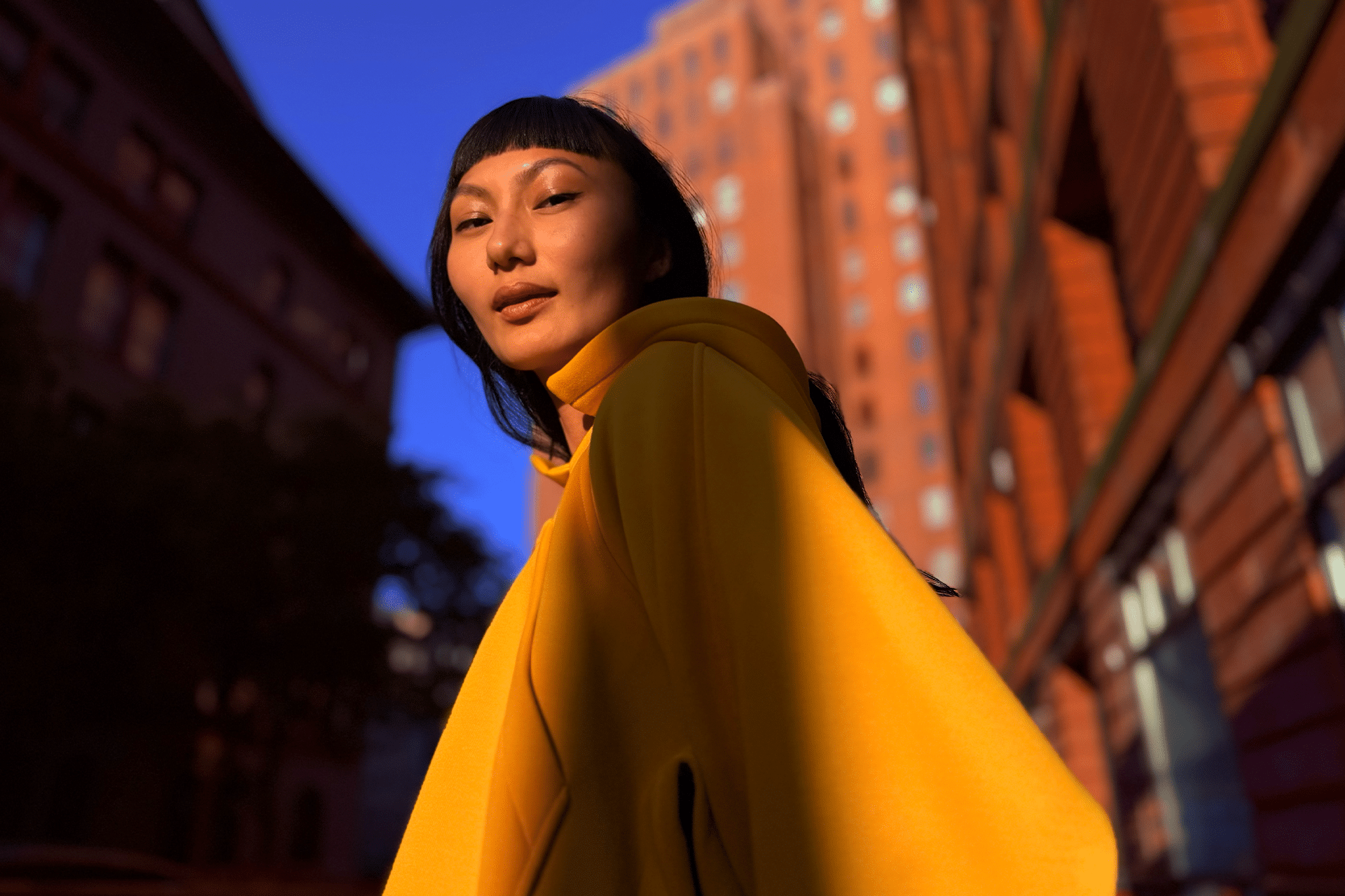 A person wearing a bright yellow coat stands on a city street, partially lit by sunlight, captured using iPhone photography. Red-brick and brown buildings rise in the background beneath a clear blue sky.