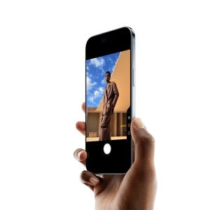A hand holds an iPhone 17 Pro displaying a photo of a person in a brown outfit standing outdoors against a blue sky and tan building, with the camera app interface visible after a recent software update.