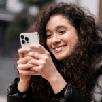 A young woman with curly hair and a black jacket smiles while looking at her smartphone outdoors, discovering new iPhone shortcuts to boost her productivity in 2025.