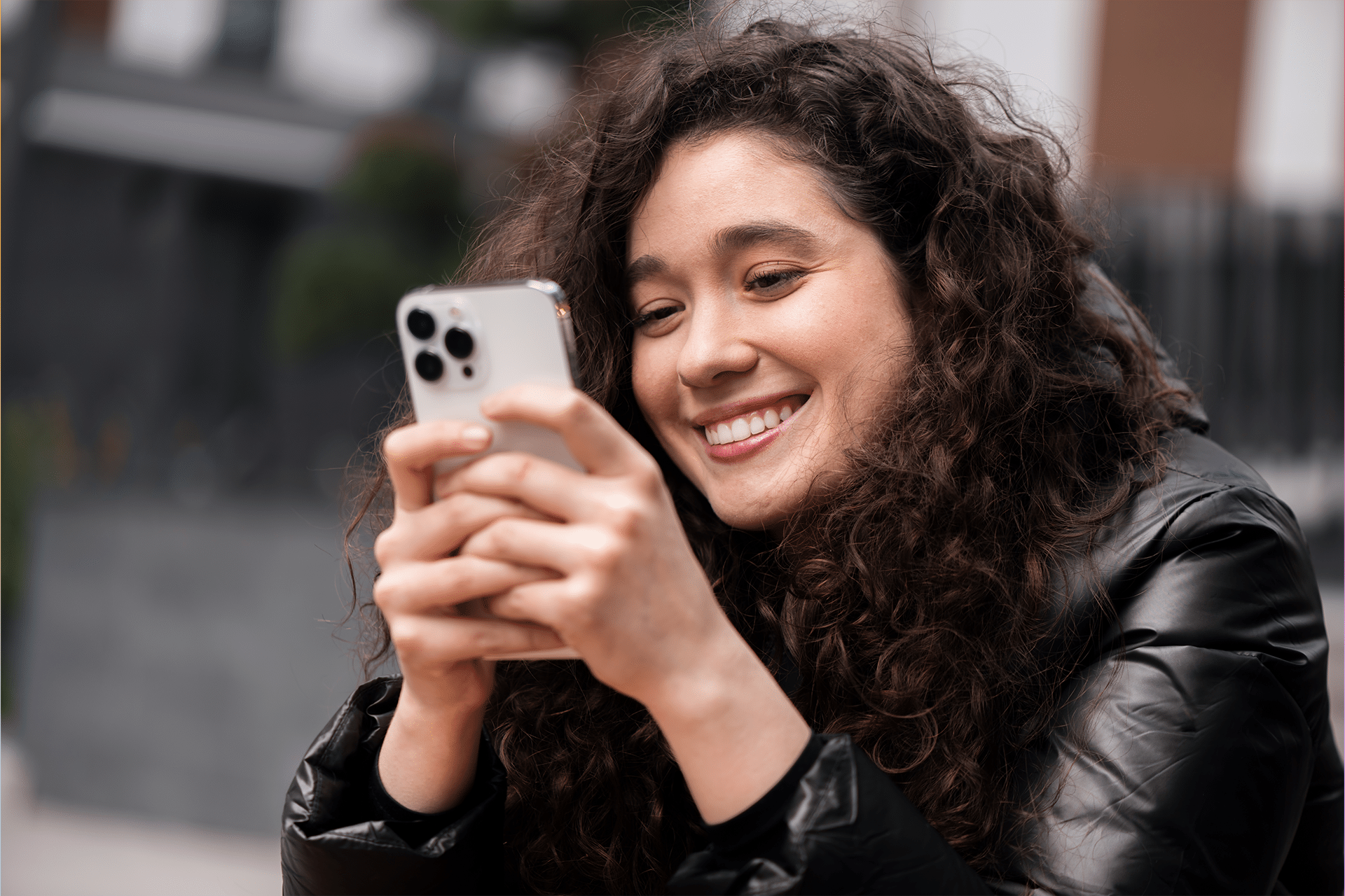 A young woman with curly hair and a black jacket smiles while looking at her smartphone outdoors, discovering new iPhone shortcuts to boost her productivity in 2025.