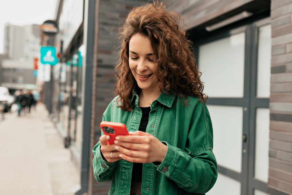 A young woman with curly hair, wearing a green jacket, stands outside near buildings and smiles while looking at her red smartphone, delighted by the new iOS 26 weather route alerts.