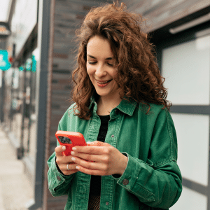 A young woman with curly hair, wearing a green jacket, stands outside near buildings and smiles while looking at her red smartphone, delighted by the new iOS 26 weather route alerts.