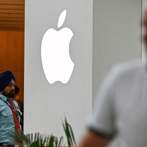 A security guard stands near a large, illuminated Apple logo and a colorful grand opening sign—an event highlighting Apple India’s expanding iPhone supply chain—as a blurred person in the foreground talks on the phone.