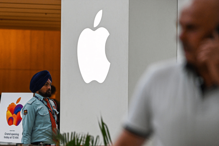 A security guard stands near a large, illuminated Apple logo and a colorful grand opening sign—an event highlighting Apple India’s expanding iPhone supply chain—as a blurred person in the foreground talks on the phone.