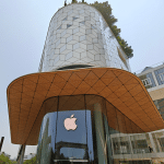 A modern Apple store with a curved glass entrance, large overhanging roof, and hexagonal-patterned upper facade, showcasing Apple India’s innovation in design amid office buildings and trees, reflecting its growing iPhone supply chain presence.