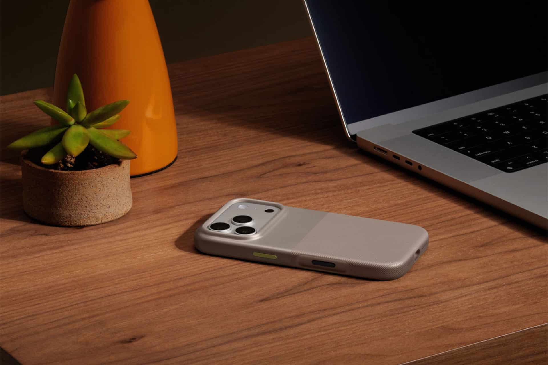A smartphone in a beige iPhone cover lies on a wooden desk beside a laptop, a small potted succulent, and an orange vase.