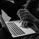 A person sits with a laptop on their lap, using one hand to type while the other is raised mid-gesture. The image, in black and white, captures the focused energy of remote workers using MacBook productivity hacks in 2025.