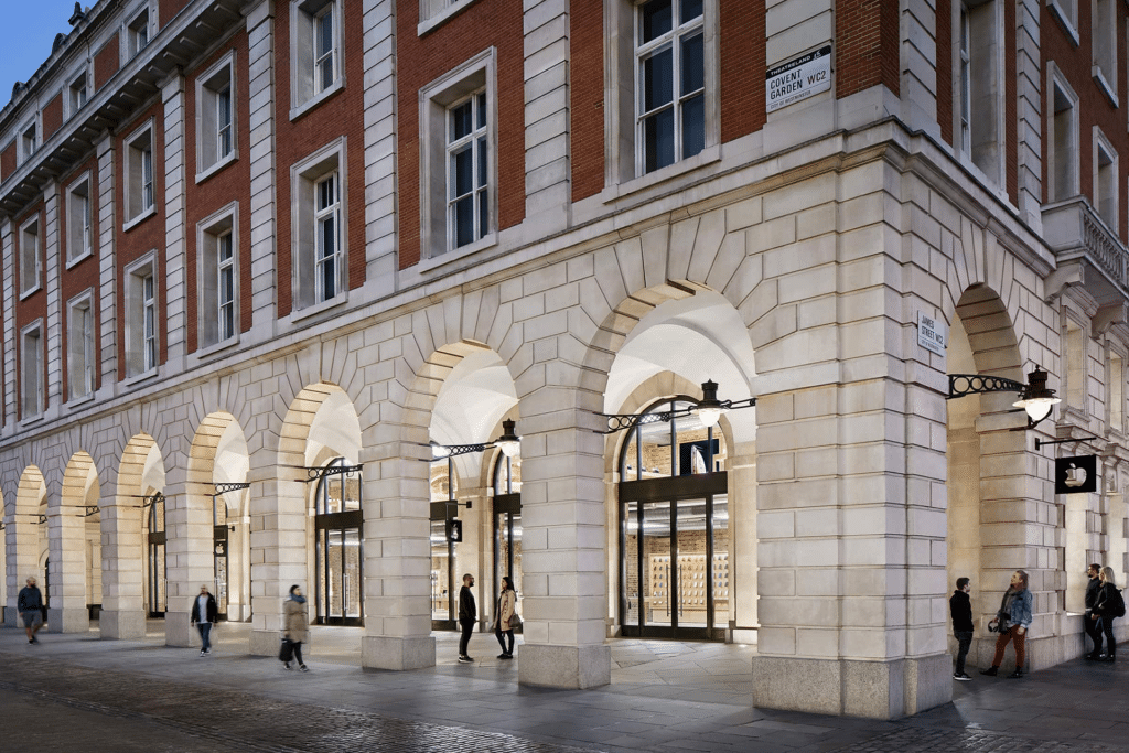 People walk past a large building with red brick upper floors and light stone arches at street level, featuring tall windows and glass doors; street lamps hang from the walls—reflecting the city's historic adherence to antitrust rules.