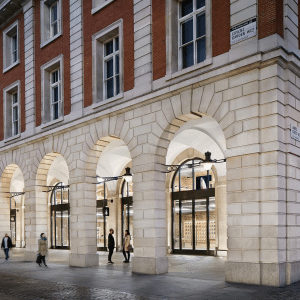 People walk past a large building with red brick upper floors and light stone arches at street level, featuring tall windows and glass doors; street lamps hang from the walls—reflecting the city's historic adherence to antitrust rules.