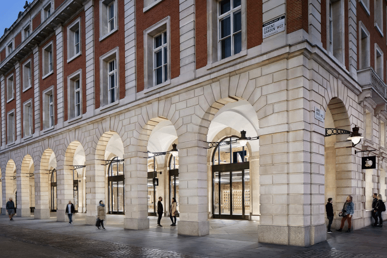 People walk past a large building with red brick upper floors and light stone arches at street level, featuring tall windows and glass doors; street lamps hang from the walls—reflecting the city's historic adherence to antitrust rules.