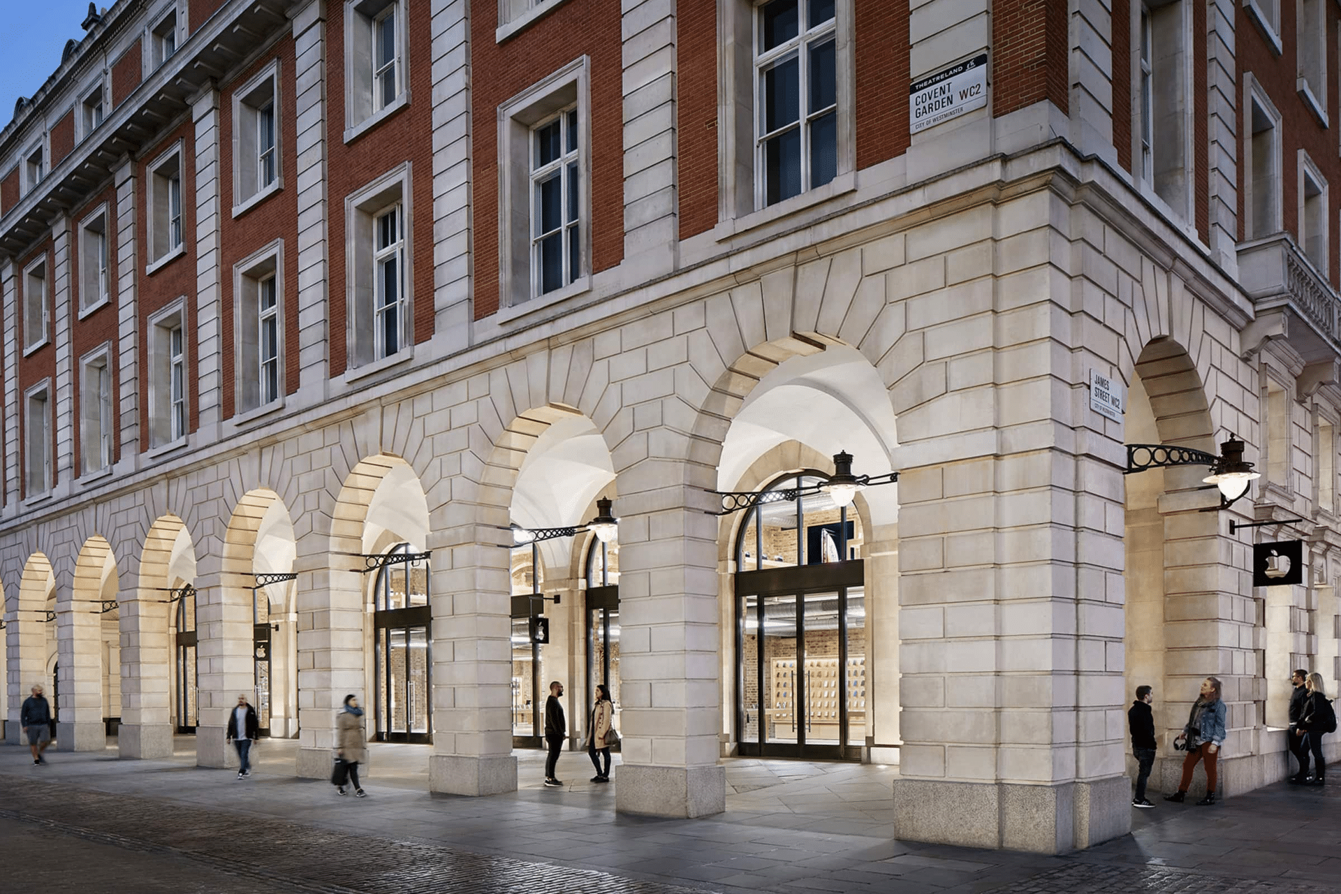 People walk past a large building with red brick upper floors and light stone arches at street level, featuring tall windows and glass doors; street lamps hang from the walls—reflecting the city's historic adherence to antitrust rules.