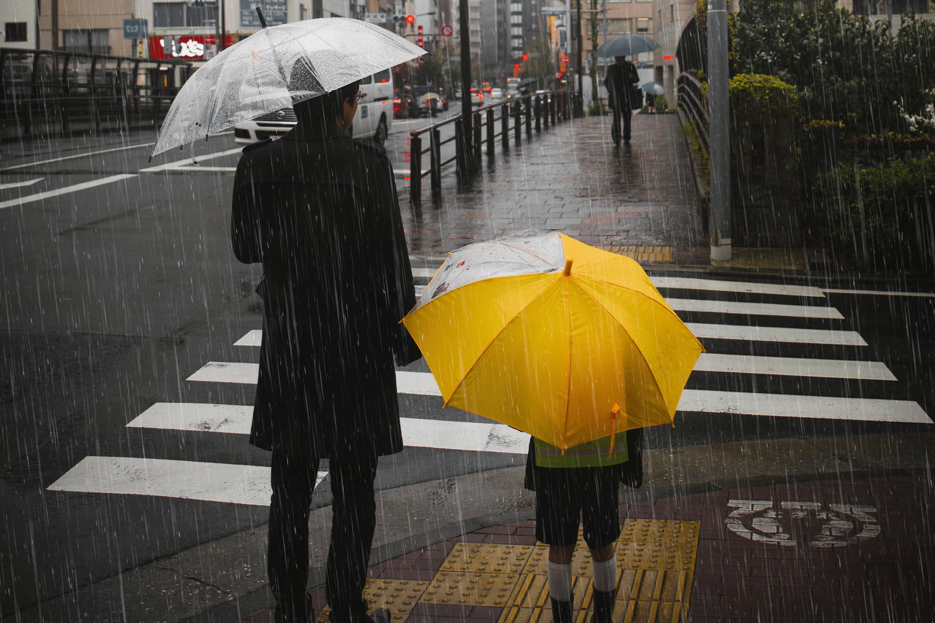 An adult and a child holding umbrellas, one clear and one bright yellow, cross a city street in the rain. Rain streaks are visible as iOS 26 weather route alerts guide their way, with the crosswalk and wet pavement reflecting the urban surroundings.