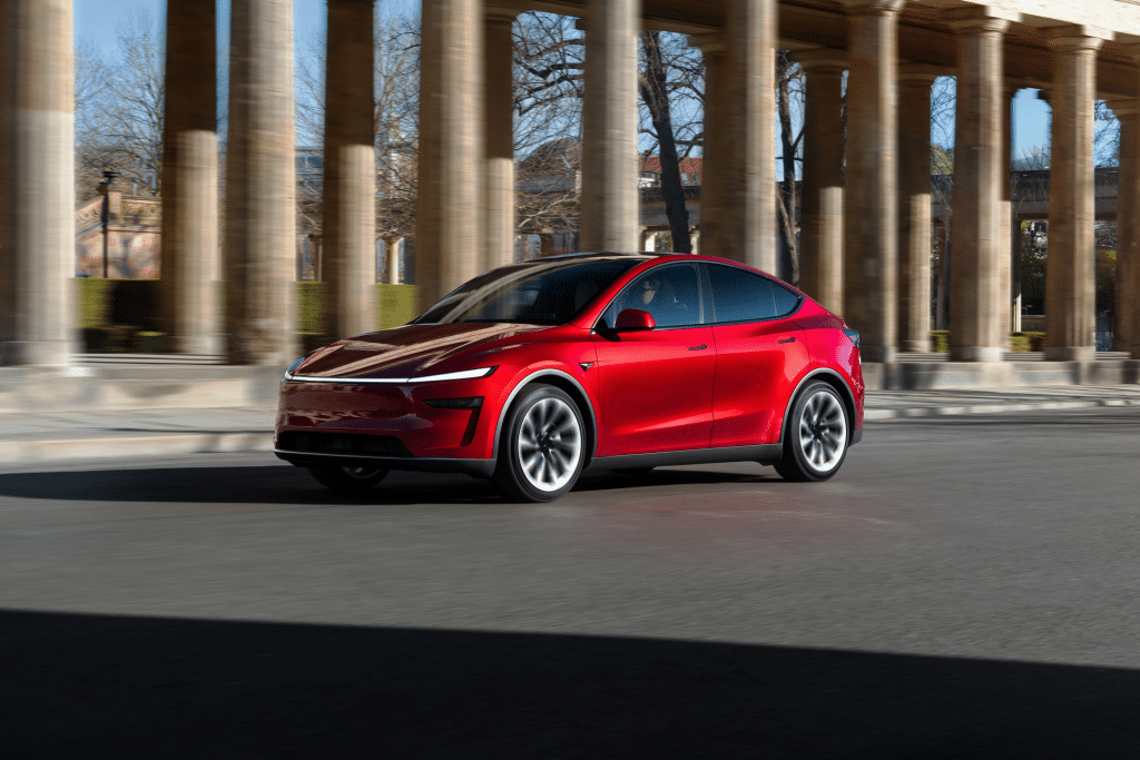 A sleek, red Tesla Model Y 2025 electric SUV drives past tall stone columns on a sunny day, showcasing its performance with trees and buildings visible in the background.