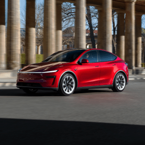 A sleek, red Tesla Model Y 2025 electric SUV drives past tall stone columns on a sunny day, showcasing its performance with trees and buildings visible in the background.
