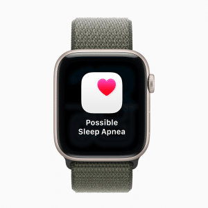 A smartwatch with a green fabric band displays a screen showing a heart icon and the text "Possible Sleep Apnea" on a black background, highlighting advanced features like hypertension alerts.