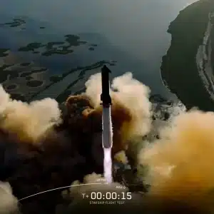 A rocket launches, surrounded by clouds of smoke, over a coastal area with water and greenery visible during the SpaceX 11th Starship test flight. On-screen data shows a speed of 146 and altitude of 0.3, with a timer at T+00:00:15.
