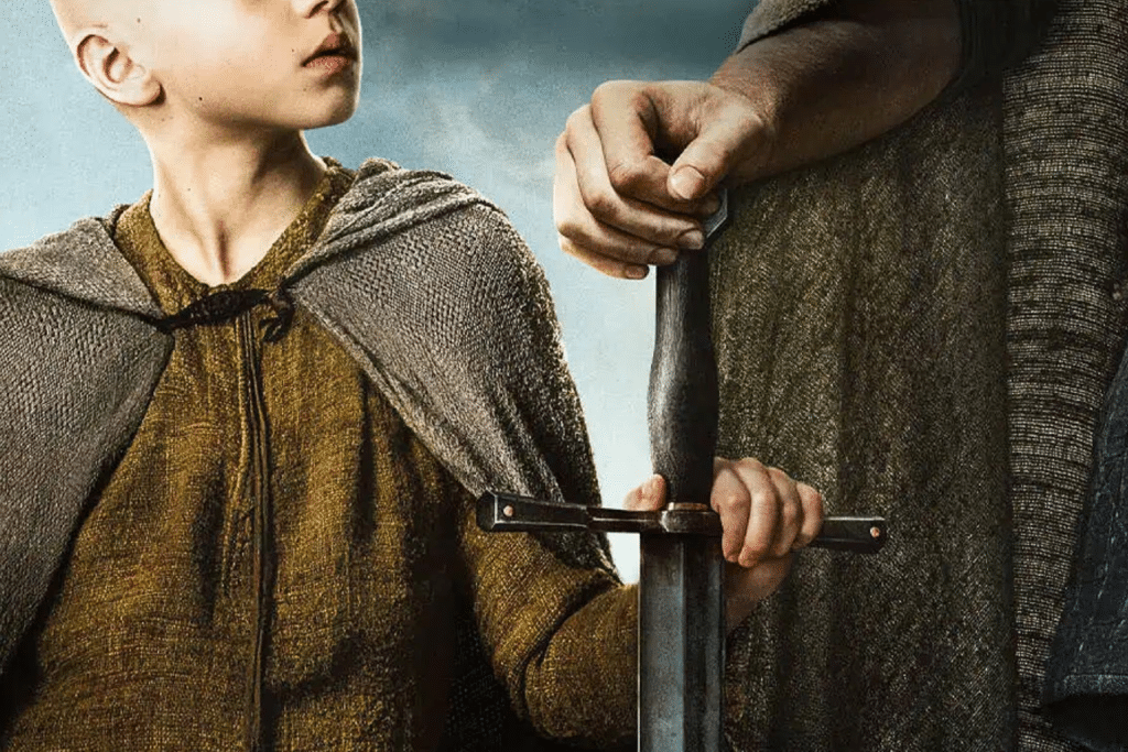 A young boy in a brown cloak looks up while holding the hilt of a large sword, shared with an adult whose upper body is mostly out of frame—a moment worthy of A Knight of the Seven Kingdoms.