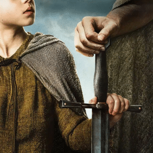 A young boy in a brown cloak looks up while holding the hilt of a large sword, shared with an adult whose upper body is mostly out of frame—a moment worthy of A Knight of the Seven Kingdoms.