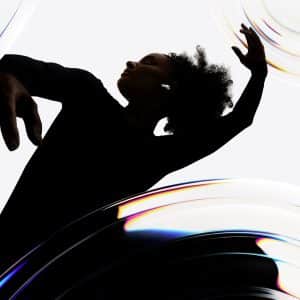 A person in a black outfit dances gracefully with arms raised, surrounded by swirling, translucent, rainbow-tinted shapes on a white background.