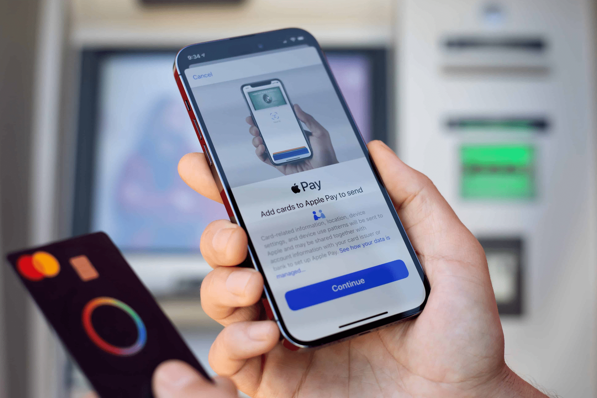 A person holds a smartphone setting up Apple Pay, with a bank card in the other hand. An ATM machine is blurred in the background, highlighting the importance of staying alert to Apple Pay scams.