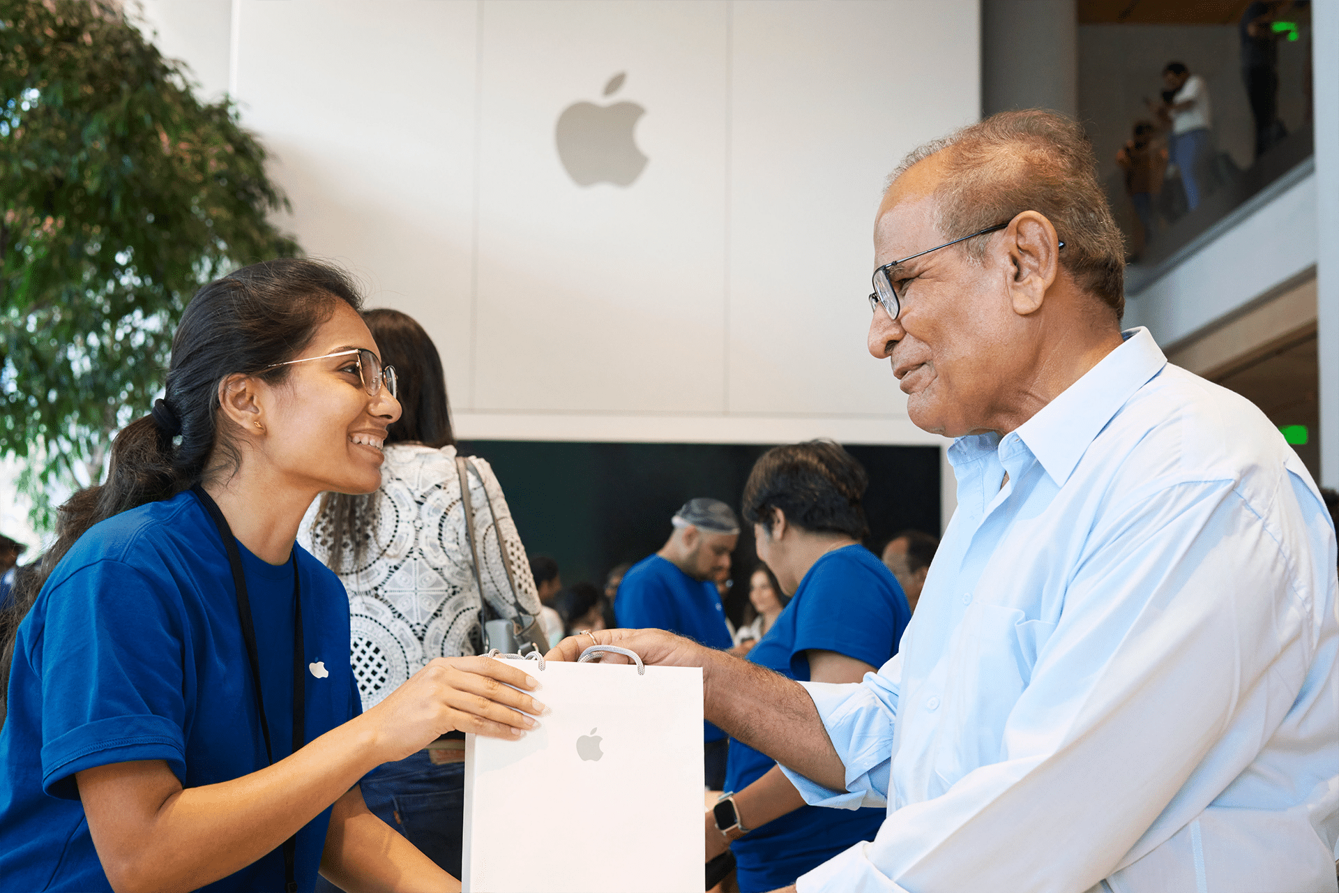 A smiling Apple Store employee hands a bag with the Apple logo to an older man in a light blue shirt. They stand inside a bright, modern Apple Store with the logo visible in the background, amid rumors about potential Apple layoffs 2026.