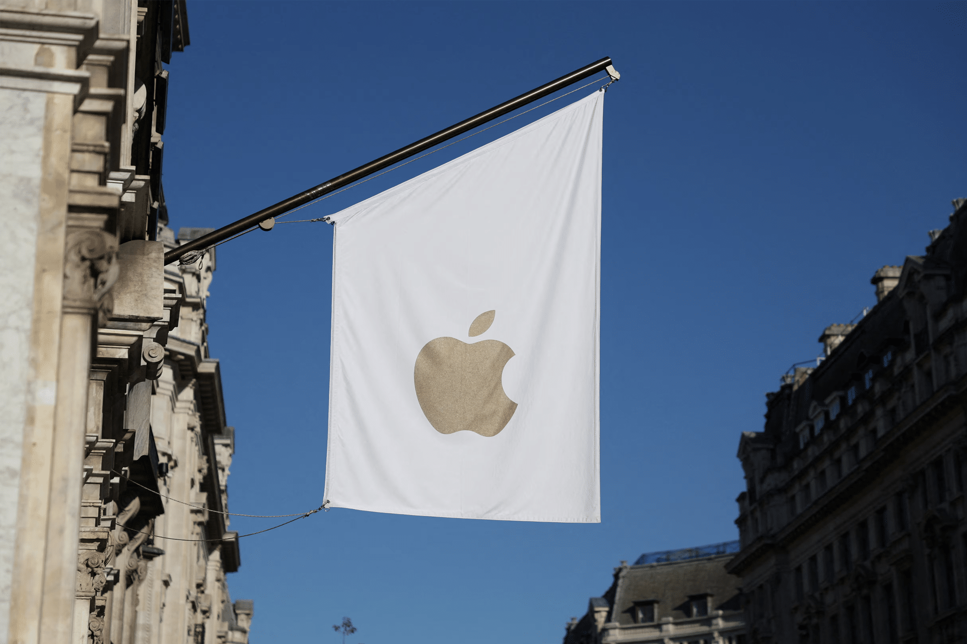 A white flag with a gold Apple logo hangs outside a building, set against a clear blue sky and surrounded by historic stone architecture, symbolizing the presence of UK Apple and its dedication to protecting encrypted iPhones.